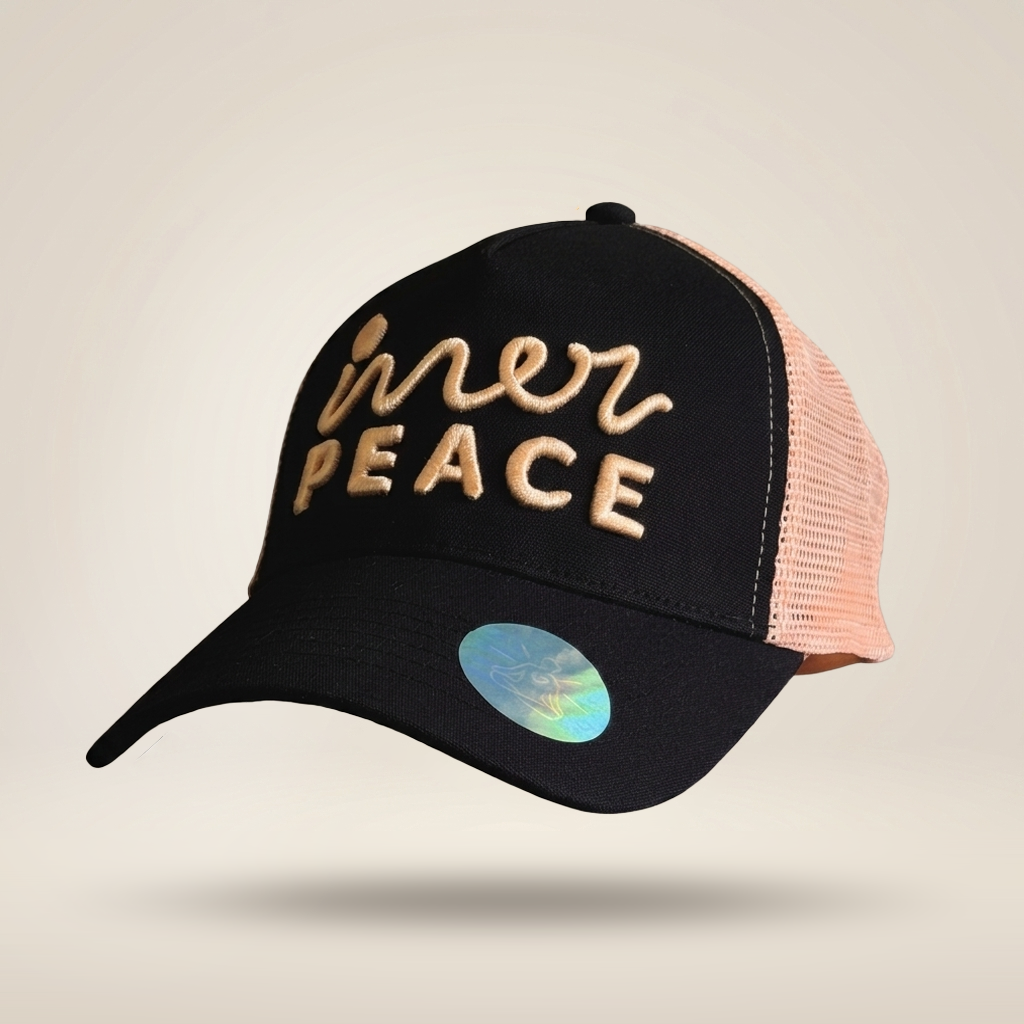 A black trucker-style cap with peach mesh back panels and gold embroidered text reading 'inner peace' on the front, shown on a light beige background.