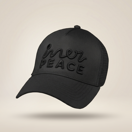 A black trucker-style cap with a curved brim, black mesh back panels, and tonal black embroidered text reading 'inner peace' on the front, shown against a light beige studio background.