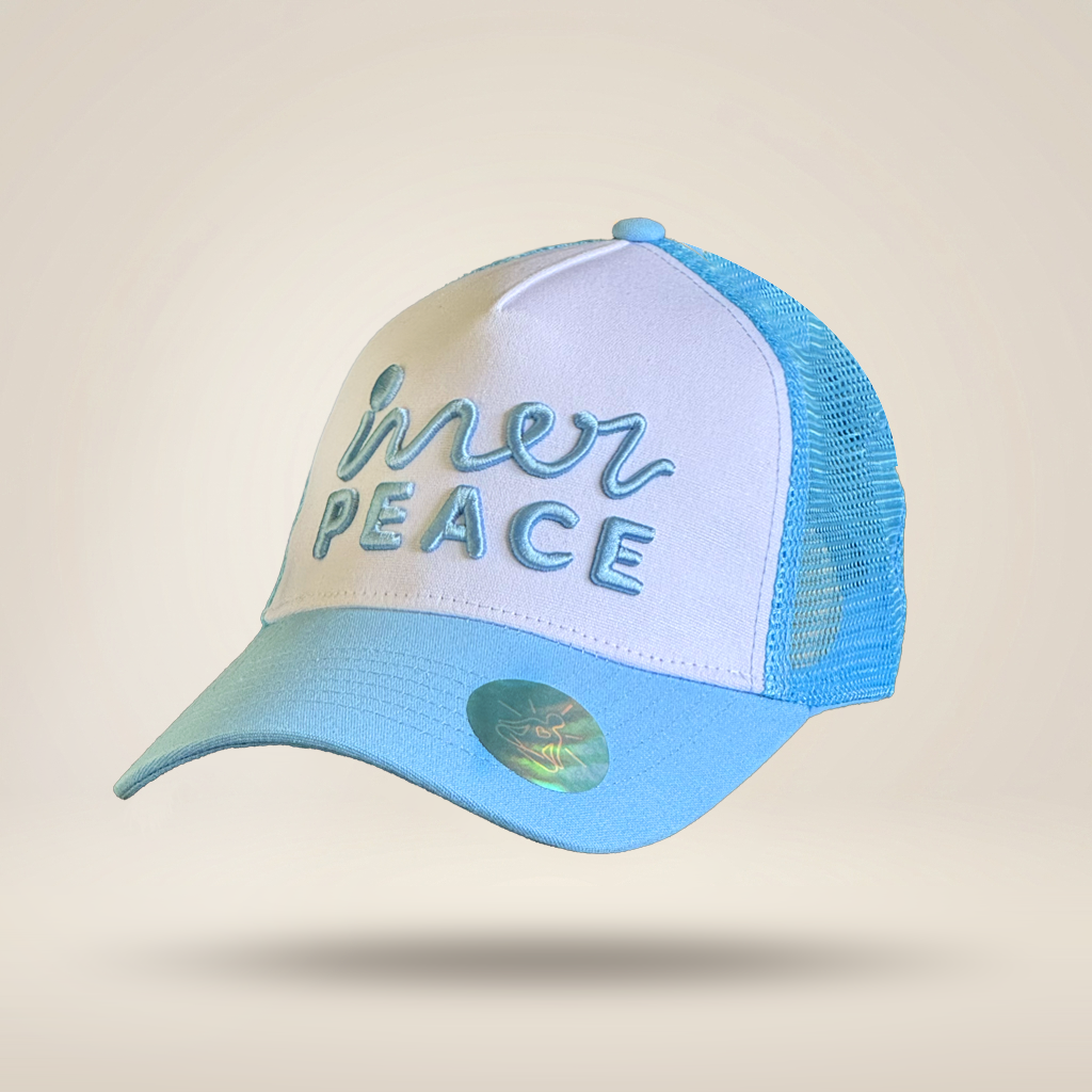 A light blue and white trucker hat with 'inner peace' embroidered on the front, shown against a solid beige background.