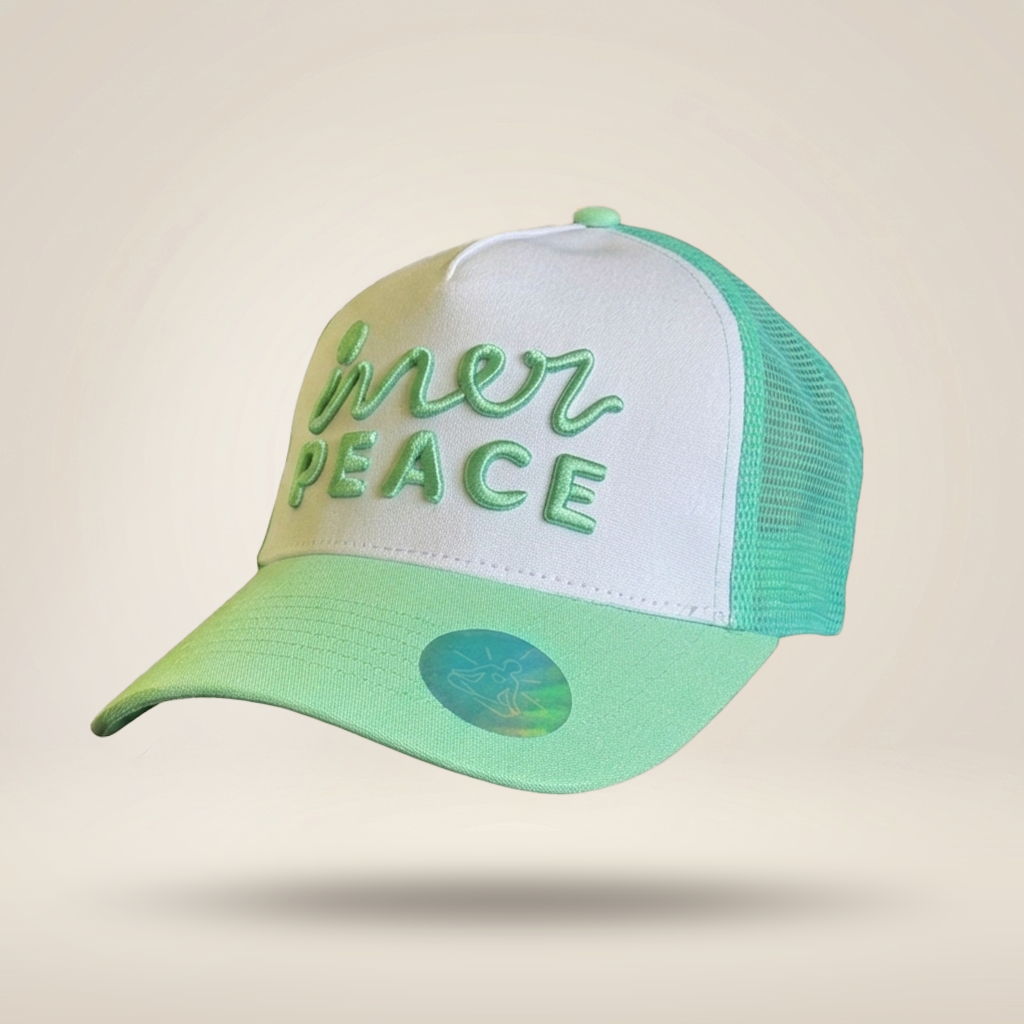 A trucker-style cap with a white front panel, mint green mesh back, and a mint green brim featuring the embroidered text 'inner peace' in green. A small holographic sticker is visible on the brim, set against a solid beige background.