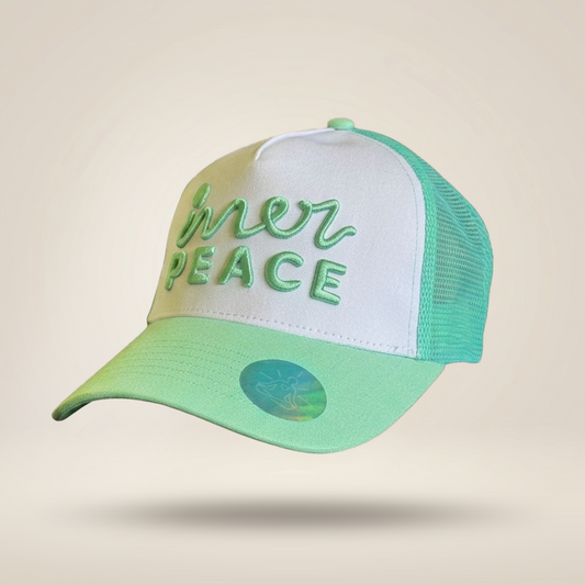A trucker-style cap with a white front panel, mint green mesh back, and a mint green brim featuring the embroidered text 'inner peace' in green. A small holographic sticker is visible on the brim, set against a solid beige background.