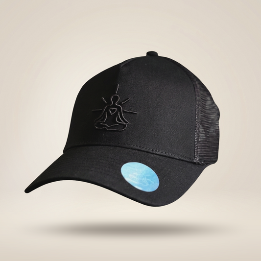 A black trucker-style baseball cap with a curved brim, black mesh back panels, and a black embroidered meditating figure logo on the front. A light blue circular sticker is visible on the brim, set against a solid beige background.