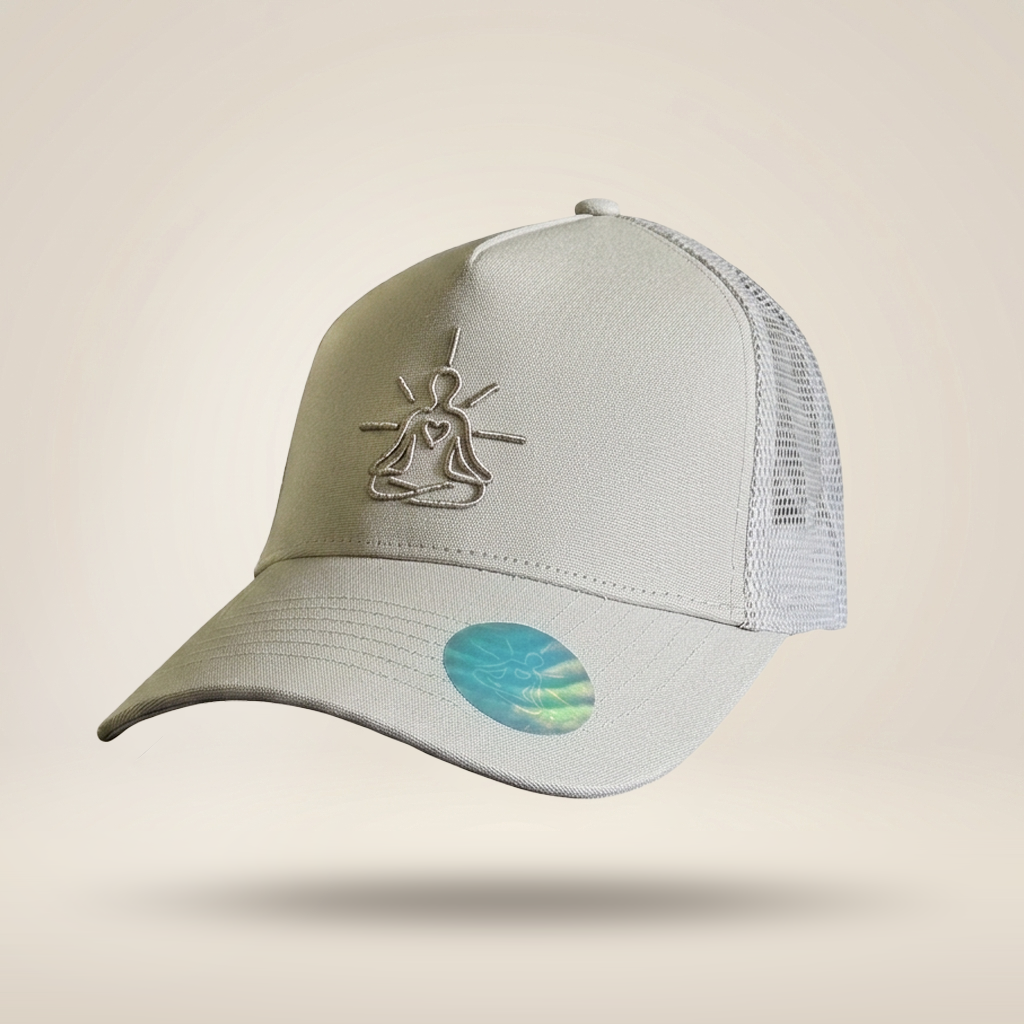 A light gray trucker-style baseball cap with a mesh back, featuring a metallic silver line-art meditating figure embroidered on the front and a holographic oval sticker on the brim, set against a solid beige background.