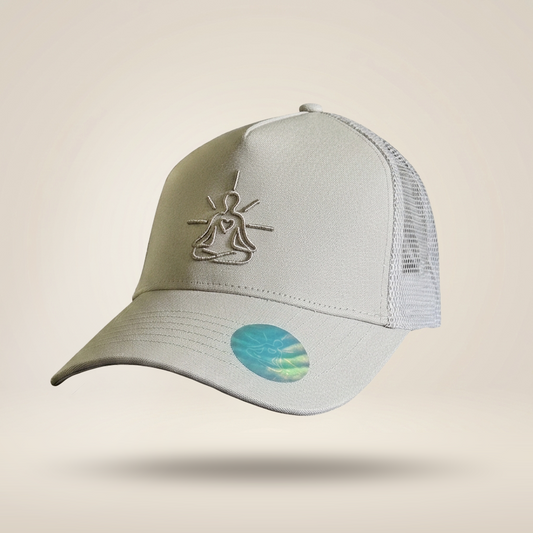 A light gray trucker-style baseball cap with a mesh back, featuring a metallic silver line-art meditating figure embroidered on the front and a holographic oval sticker on the brim, set against a solid beige background.