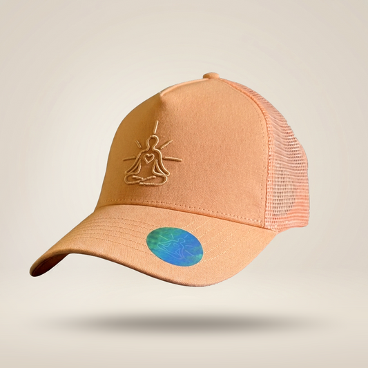 A peach-colored trucker hat with a mesh back, featuring an embroidered meditating figure logo on the front and a holographic sticker on the brim, set against a light beige background.